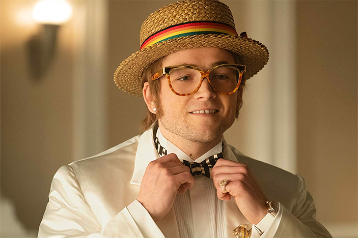 Taron Egerton as Elton John in Rocketman, wearing a straw hat and adjusting his bowtie