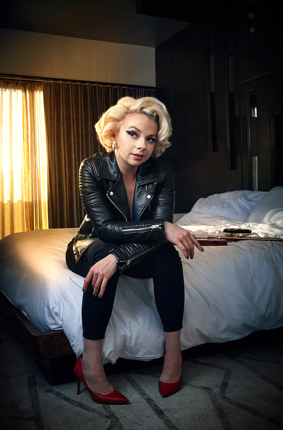 Samantha Fish  with her guitar sitting on a bed in a black outfit with red high heels